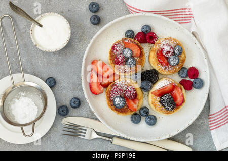 Quark Pfannkuchen, syrniki, Quark Krapfen mit frischen Beeren (Himbeere, Erdbeere, Heidelbeere, BlackBerry) und Puderzucker in eine weiße Platte. Stockfoto