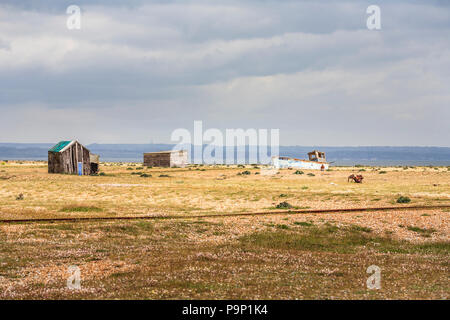 Alte Holzhütten und einem verlassenen Fischerboot auf der Kiesstrand in Dungeness, Shepway District, Kent mit einem bewölkten grauen Himmel Stockfoto