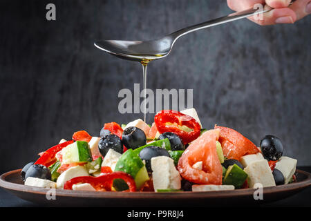 Woman's Hand gießt ein Griechischer Salat mit Olivenöl aus einem Löffel auf dunklem Hintergrund Stockfoto