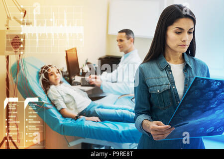 Auf EEG-Ergebnisse. Ruhe aufmerksame junge Mutter mit Blick auf die EEG-Ergebnisse ihrer Tochter beim Stehen nicht weit von ihr in einer modernen Klinik Stockfoto