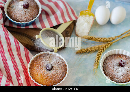 Kuchen mit cerries auf einem Tisch in kleinen Porzellan Tassen. Es gibt Eier, Mehl, Weizen, Puderzucker neben Kuchen. Stockfoto