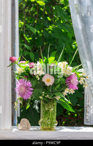 Ein Blumenstrauß von Clover, Kornblumen und Jasmin in einem Glas Vase und ein Herz aus Stein auf der Fensterbank eines offenen Fensters eines Dorfes Haus Stockfoto