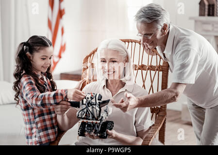 Es betrachten. Lächelnd Oma sitzt im Schaukelstuhl, die Roboter während glücklich und Opa neben ihr und berührt, Spielzeug stehend Stockfoto