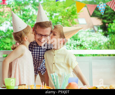 Zwei süße Kinder küssen ihre junge und glückliche Vater bei seiner Geburtstagsfeier auf bunt dekorierten Terrasse zu Hause Stockfoto