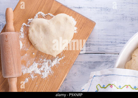 Teig für hausgemachte Kuchen, heart-shaped, auf einem Holzbrett und rollen Sie ein Handtuch mit einem Muster auf weißem Hintergrund. Freier Platz für Text oder Werbung Stockfoto