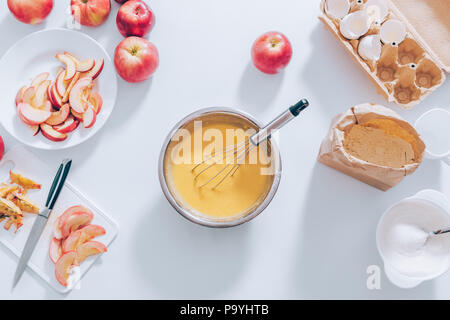 Zutaten und Werkzeuge für das Kochen Apfelkuchen, Ansicht von oben. Metallschale mit Teig, Schneebesen gefüllt, geschnittenes Obst, Eierschalen, Mehl und Zucker, Flachbild Composit legen Stockfoto