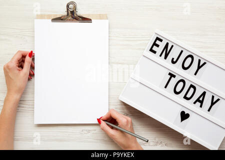 Weibliche Hände mit einem leeren noticepad und genießen heute' Wort auf Lightbox über weiß Holz- Hintergrund, Ansicht von oben. Von oben, flach und Overhead. Stockfoto