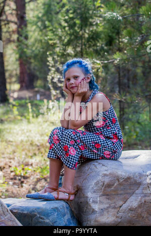 Atrtractive 8 jähriges Mädchen, in bunten jump Suit, die Haare hell blau gefärbt, sitzen auf den Felsen, mit Blick auf die Kamera, den Kopf auf die Hände. Model Release Stockfoto