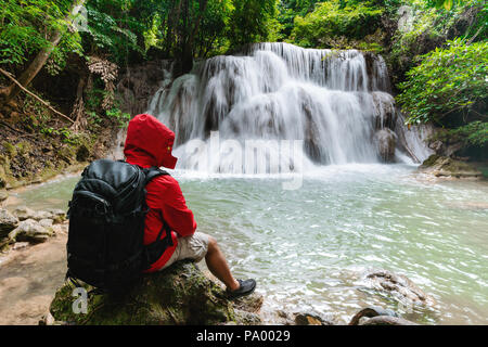 Ein Mann im roten Mantel mit Rucksack auf dem Felsen genießen Sie einen wunderschönen Wasserfall in tropischen Regenwald Huay Mae Khamin in Kanchanaburi, Thailand sitzen Stockfoto