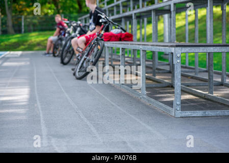 Radfahrer ruhen und sitzen auf Metall Bank neben outdoor Basketball Court und das Spiel beobachten. Gesunder Lebensstil und Outdoor-Aktivitäten in Summ Stockfoto