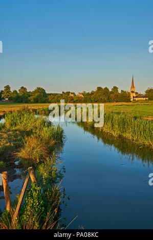 Der Fluss Windrush Wicklung durch Wasser Wiesen in Richtung der Cotswold Stadt Burford, mit dem Kirchturm von St. Johannes der Täufer in der Ferne. Stockfoto