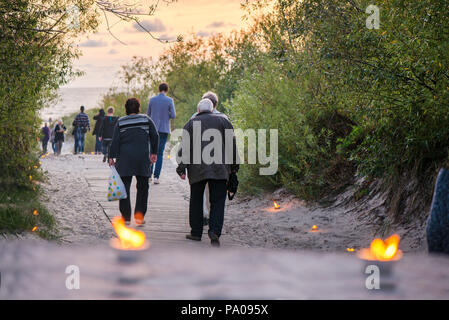 Romantische Lagerfeuer Nacht am Meer bei Sonnenuntergang. Leute versammeln um Nacht der alten Leuchten feiern. Alte Paar gehen auf Holz- Weg Stockfoto