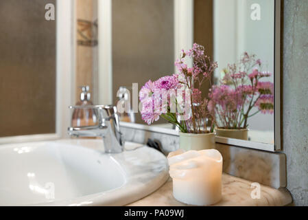Bunte Kerzenlicht neben einem modernen, gefliesten Badezimmer Waschbecken Wasserhahn eine schöne Atmosphäre. Getrocknete Blumen in Vase auf Waschbecken neben Spiegel Stockfoto