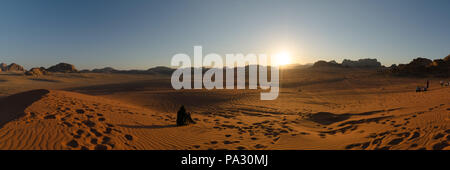 Ein weiblicher Tourist sitzt auf einer Sanddüne, die der Sonne zugewandt ist, die über Felsformationen im beliebten Trekkking-Spot Wadi Rum Desert, Jordanien, liegt Stockfoto