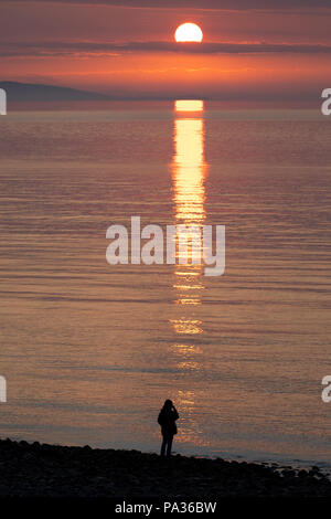 Eine einsame weibliche gegen einen schönen Sonnenuntergang und Reflexion über die Irische See bei Penmon Punkt, Anglesey, Wales Silhouette Stockfoto