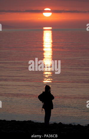 Eine einsame weibliche gegen einen schönen Sonnenuntergang und Reflexion über die Irische See bei Penmon Punkt, Anglesey, Wales Silhouette Stockfoto