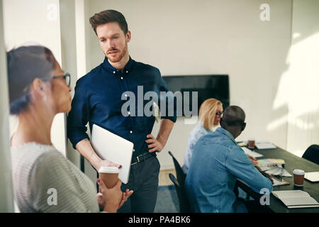 Zwei verschiedene junge Kolleginnen und Kollegen sprechen zusammen in einem Büro Boardroom mit Kollegen an einem Tisch im Hintergrund arbeiten Stockfoto