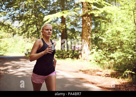 Passen junge blonde Frau in Sportkleidung alleine laufen entlang einer von Bäumen gesäumten Pfad durch den Wald an einem sonnigen Tag Stockfoto