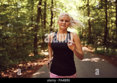 Passen junge blonde Frau in Sportkleidung lächelnd während läuft allein auf einem Pfad durch einen Wald Stockfoto