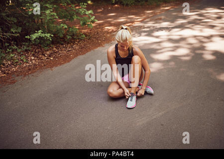 Passen junge blonde Frau in Sportkleidung sitzt alleine auf der Landstraße bis Band ihre Laufschuhe, bevor Sie für einen Lauf Stockfoto