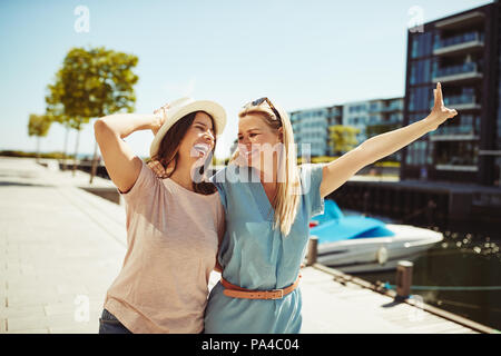 Zwei unbeschwerte Junge weibliche Freunde lachen beim Gehen Arm in Arm zusammen in der Stadt im Sommer Stockfoto