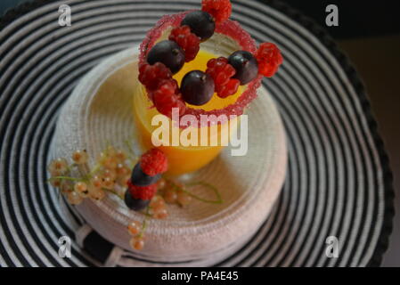 Glas mit Orangensaft, kirschrotem Zuckerrand und Früchten darauf, Himbeeren, Stachelbeeren und Johannisbeeren auf einem weiblichen Hut. Stockfoto