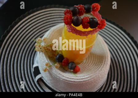 Glas mit Orangensaft, kirschrotem Zuckerrand und Früchten darauf, Himbeeren, Stachelbeeren und Johannisbeeren auf einem weiblichen Hut. Stockfoto