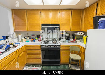 Unordentliche alte Wohnung Küche mit Eiche Schränke, Arbeitsplatten Fliesen, Gasherd, grüne Bodenbeläge und Stapel von Gerichten. Stockfoto