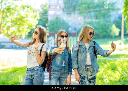 Drei Mädchen im Teenageralter. Sommer in der Natur. In seinen Händen hält ein Smartphone. Nehmen Sie Fotos auf dem Telefon. Bei Sonnenbrillen und Denim Kleidung. Das Konzept der Selbst Stockfoto