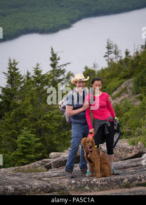 Cowboy Hut tragen junge Mann mit asiatischen Frau und Golden Retriever Welpen für Foto mit Pinien und grossen See im Hintergrund posiert. Stockfoto