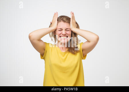 Schreckliche Kopfschmerzen. Frustrierte europäische junge Frau im gelben t-hirt berühren ihren Kopf mit Händen und Gesicht voller Schmerz, während gegen Ständigen Stockfoto