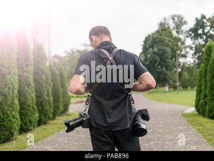 Zurück professioneller Hochzeitsfotograf in schwarzen T-Shirt und mit zwei Kameras, ein Schulterriemen gegen die grünen Garten. Hochzeit Fotographie. Stockfoto