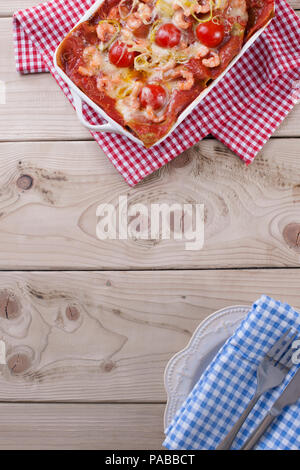 Lasagne gebacken in einem weißen Backblech, auf eine Serviette in einem roten Käfig und ein Teller mit einem blau serviette auf einem hölzernen Hintergrund Stockfoto
