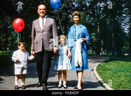 Familie mit drei Generationen in einem New York City Park, USA in den 1960er Jahren. Zur Familiengruppe gehören ein Großvater, eine junge Frau und eine Mutter, ein 2-jähriger Junge und ein vierjähriges Mädchen. Die Kinder halten bunte Luftballons. Die Familie ist in Sonntagskleidung gekleidet. Der kleine Junge trägt einen Seersucker-Anzug mit Shorts. Das kleine Mädchen trägt ein Partykleid Stockfoto