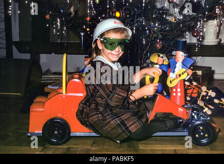 Junge 4-jährige Mädchen posiert vor einem Weihnachtsbaum in einem Wohnzimmer am Weihnachtstag in den USA in den 960er Jahren. Sie lächelt und trägt ein Tartan-Kleid, einen Helm und eine Brille, sitzt in einem Spielzeugpedal Rennwagen. Unter dem Weihnachtsbaum steht ein Tinkertoy-Bauset Stockfoto