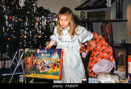 Verschlafene aussehende junge vierjährige Mädchen mit langen blonden Haaren trägt ein Nachthemd Auspacken ein Weihnachtsgeschenk vor einem Weihnachtsbaum in einem Wohnzimmer am Weihnachtsmorgen in den 1960er Jahren, USA. Das Geschenkpapier wird beiseite geworfen. Das Weihnachtsgeschenk ist eine große Box namens Walt Disney's Sleeping Beauty Coloring Fun Stockfoto