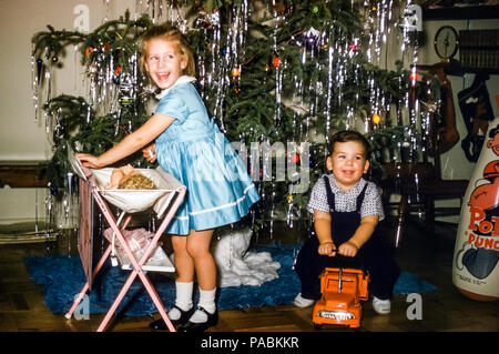 Junge dreijährige Mädchen und ein Jahr alten Jungen Kleinkind vor geschmückten Weihnachtsbaum spielen mit Spielzeug in den 1950er Jahren. Das lächelnde Mädchen trägt ein blaues Kleid und spielt mit einer Puppe in einem Spielzeugbecken. Der kleine Junge lacht und sitzt auf einem Spielzeug-Kipper neben einem Popeye-Boxsack Stockfoto