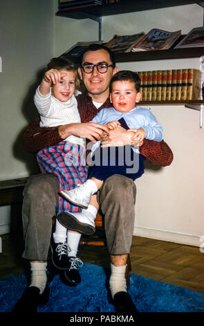 Familie mit Vater und zwei kleinen Kindern, einem kleinen Mädchen und einem Kleinkind, die in den 1950er Jahren in einem Wohnzimmer mit Bücherregalen mit Büchern und Zeitschriften in den USA auf dem Schoß saßen Stockfoto