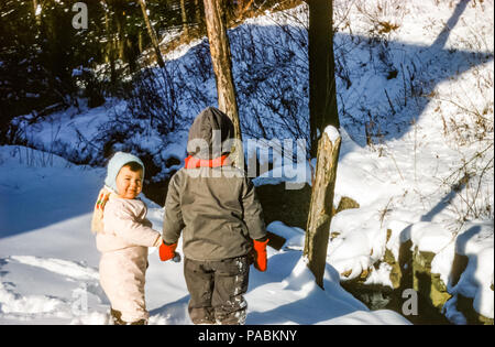 Kleine Kinder, Bruder und Schwester halten sich die Hände, tragen Winterschneesachen im Schnee in Sonnenschein, USA in den 1950er Jahren Stockfoto