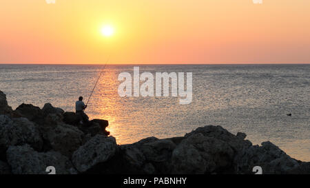 Ein einsamer Angler auf den Felsen am Mandraki Hafen in Rhodos, Griechenland bei Sonnenaufgang. Stockfoto