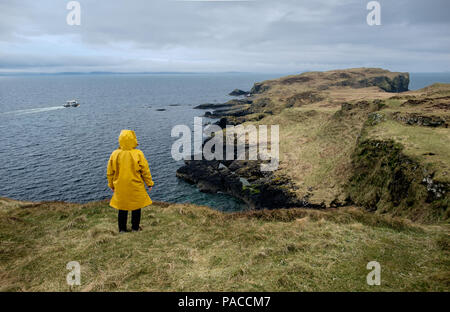 Frau mit Gelb oilskin Jacke beobachten ein Boot von Staffa Insel, in der Nähe der Isle of Mull, Schottland Stockfoto