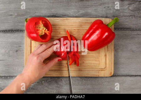 Table Top - Frau Hände, schneiden Red Paprika mit Chefs Messer auf altem Schneidebrett, mit grauen Holz Schreibtisch unter. Stockfoto
