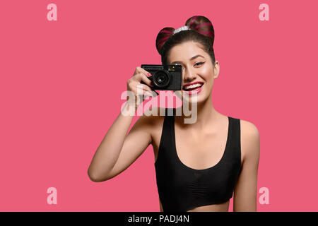 Vorderansicht der lächelnden, glücklichen Mädchen unter Foto mit alten Kamera. Lachen, Kamera, in ausgefallenen Frisur mit einem Bogen, bunten Tag bilden mit rosa Lippenstift. Posieren auf einem hellen Rosa Hintergrund. Stockfoto