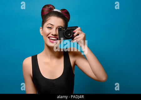 Lächelnd, glücklich, Foto mit alten Kamera. Lachen, Kamera, in ausgefallenen Frisur mit einem Bogen, bunten Tag bilden mit rosa Lippenstift. Auf gesättigte tief blauen Hintergrund. Stockfoto