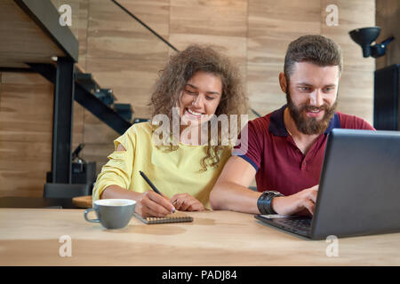 Studium Studenten in gemütlicher Café sitzen, mit Laptop, Lächeln, Lachen, trinken Kaffee, Studieren, Schreiben von Notizen. Auf Holztisch mit schwarz metallic Treppen für den Hintergrund. Stockfoto