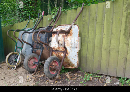 Drei (3) rostige alte Schubkarren lehnte sich gegen ein verwittertes Holz Garten Zaun mit dem Grün im Hintergrund Stockfoto