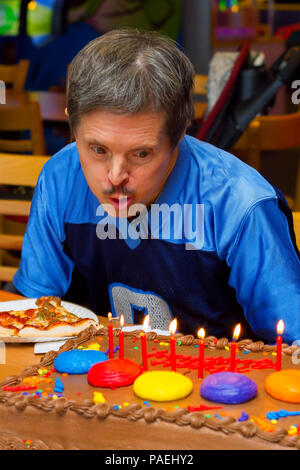 Ein älterer Mann mit Down-syndrom bläst die Kerzen auf dem Geburtstagskuchen an einer Pizza Party in seiner Ehre. Er sieht sehr konzentriert, wie er versucht zu löschen. Stockfoto