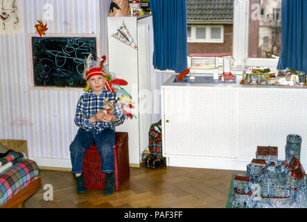 Der Junge trägt einen indianischen Kopfschmuck und sitzt in einem Kinderzimmer mit einem Kuscheltier mit Spielzeug, darunter eine Texaco-Garage und ein deutsches Elastolin-Hausser-Plastikspielzeug mittelalterliche Burg Schaffhausen Knights Castle, London, England, Großbritannien in den 1960er Jahren Stockfoto