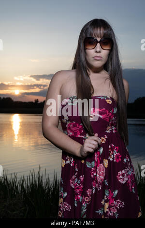 Spanisch-amerikanischen weiblichen stehen am Rande eines Sees in Cambridgeshire. Das Tragen einer Pflaume farbige geblümten Kleid und Sonnenbrille während ein Sommer Sonnenuntergang. Stockfoto