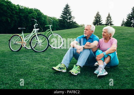 Positive alte Paar umarmen, sitzt auf dem Gras, mit ihren Fahrrädern Stockfoto